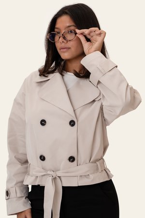 Frau, die ihre runde Brille mit Schildpattfassung zurechtrückt und einen beigen, doppelt geknöpften Trenchcoat mit schwarzen Knöpfen und gebundenem Taillengürtel trägt.