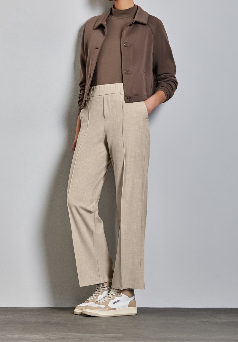 Personne portant un pantalon beige à jambes larges, une veste courte marron sur un haut marron, et des baskets beige et blanches, debout contre un mur clair.