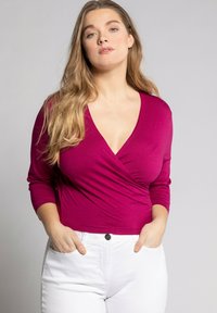 Fuchsiový top s hlubokým výstřihem do V a dlouhými rukávy, kombinovaný s bílými džíny s vysokým pasem. Tkanina se zdá být měkká a hladká.
