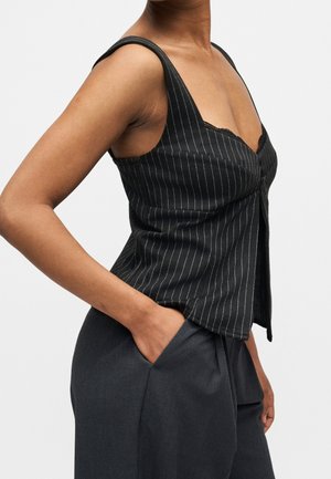 Femme portant un haut sans manches noir à fines rayures avec boutons et un pantalon gris taille haute, main dans la poche, sur un fond uni.