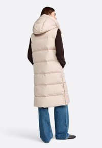 Beige puffer vest with a hood, featuring a quilted design, zipper details, and a long silhouette, paired with blue wide-leg jeans.
