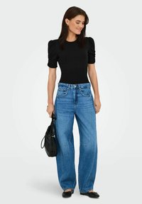 Bodycon nero a coste con maniche a palloncino, abbinato a jeans blu a vita alta e gamba larga. La modella tiene una borsa nera e indossa scarpe nere flat.