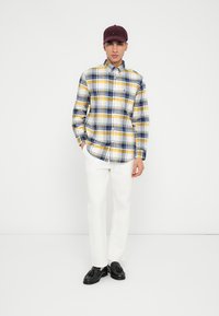 Camisa de cuadros en azul, amarillo y blanco, combinada con pantalones blancos y zapatos negros, complementada con una gorra burdeos.