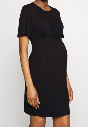 Kobieta w ciąży ubrana w czarną sukienkę z krótkim rękawem z marszczonym węzłem z przodu na wysokości talii.