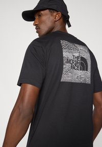 Μαύρο μπλουζάκι με γραφικό σχέδιο που περιλαμβάνει σπειροειδή μοτίβα, ριγέ διακοσμητικά και το λογότυπο "The North Face" στο πίσω μέρος. Κοντά μανίκια.