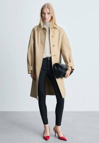 Beige trench coat s knoflíkovým zapínáním, nošený přes světle šedý svetr a černé skinny džíny. Doplněno černou kabelkou a červenými lodičkami.
