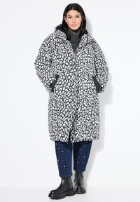 Abrigo acolchado de leopardo en blanco y negro de gran tamaño con capucha, que presenta bolsillos laterales y cierre de cremallera. Lleva puesto sobre un suéter de cuello alto gris y pantalones vaqueros.