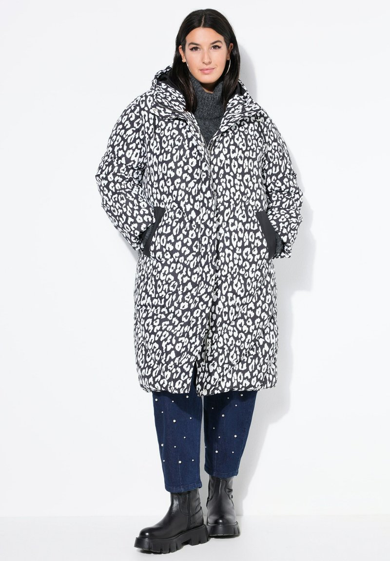 Abrigo acolchado de leopardo en blanco y negro de gran tamaño con capucha, que presenta bolsillos laterales y cierre de cremallera. Lleva puesto sobre un suéter de cuello alto gris y pantalones vaqueros.