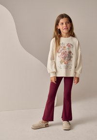 Sweatshirt crem cu grafică florală și textul "Ești magie", asortat cu pantaloni evazați vișinii și pantofi slip-on bej, pe un fundal neutru.