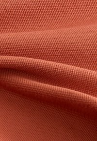 Tejido texturizado en un suave y apagado color terracota, que presenta un patrón tejido con pliegues que crean una apariencia lisa y caída.