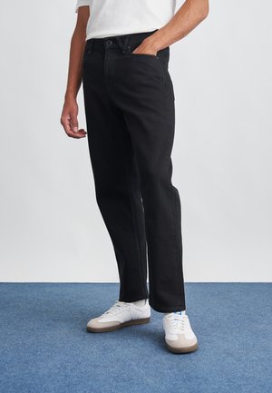 Man die zwarte spijkerbroek, witte sneakers en een wit shirt draagt, met één hand in de zak, staand op een vloer met blauw tapijt.