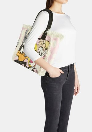 Donna che indossa un top bianco e jeans neri, che porta una borsa rosa con manici neri con personaggi dei cartoni animati e bordi frangiati.