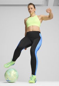 Zeleni sportski grudnjak, crne tajice s plavim naglascima i svijetlozeleni nogometni štucni. Udaranje svijetlo obojenog nogometnog lopte. Glatka tekstura.