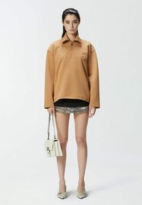 Felpa marrone oversize con zip e logo in rilievo, abbinata a una mini gonna nera con rifiniture argentate, mentre tiene in mano una borsa bianca.