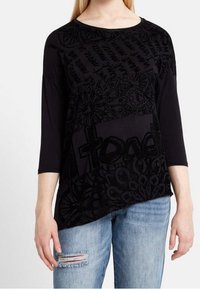 Kvinna iklädd en svart långärmad topp med abstrakta mönster och text, kombinerad med ljusblå, slitna jeans, stående framför en enfärgad bakgrund.