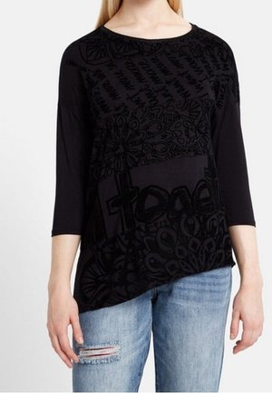 Femme portant un haut noir à manches longues avec des motifs abstraits et du texte, associé à un jean déchiré bleu clair, se tenant devant un fond uni.