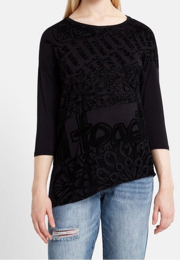 Kvinna iklädd en svart långärmad topp med abstrakta mönster och text, kombinerad med ljusblå, slitna jeans, stående framför en enfärgad bakgrund.