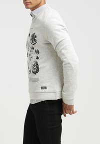 Sudadera gris claro con estampados botánicos negros en la parte delantera. Material suave con puños y dobladillo acanalados. Combinada con una camisa de cuello.