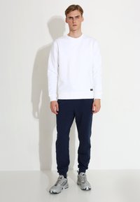 Bjela dukserica s okruglim izrezom, rebrastim manžetama i donjim rubom, u kombinaciji s tamnoplavim trenirkama. Model nosi sive tenisice i stoji pred jednobojnom pozadinom.