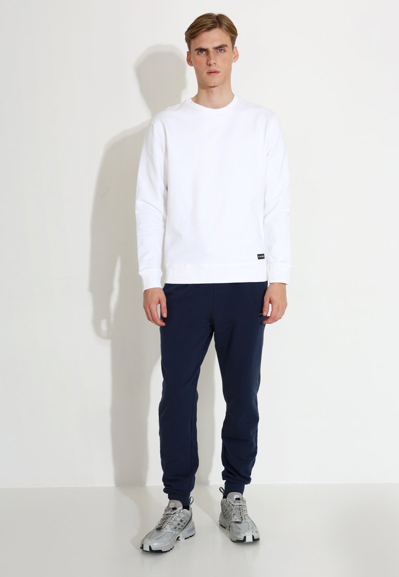 Bjela dukserica s okruglim izrezom, rebrastim manžetama i donjim rubom, u kombinaciji s tamnoplavim trenirkama. Model nosi sive tenisice i stoji pred jednobojnom pozadinom.