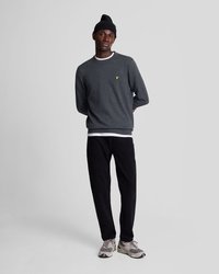 Sweatshirt gris à col rond et petit logo jaune, associé à un pantalon noir et des baskets grises. Le modèle porte également un bonnet noir.