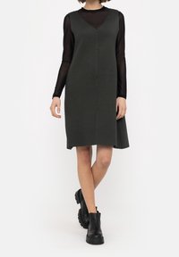 Robe sans manches vert foncé avec un col en V, fabriquée dans un tissu lisse. Associée à un haut à manches longues noir et des bottines noires épaisses.