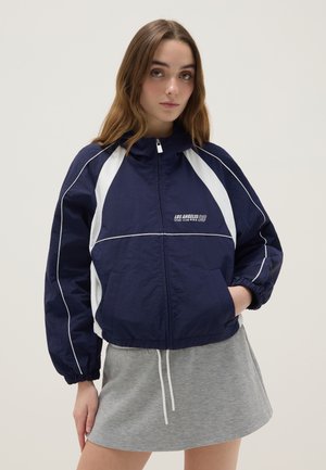 Giovane donna con giacca a vento sportiva blu navy e bianca con la scritta "Los Angeles 98", abbinata a una gonna casual grigio chiaro, con le mani nelle tasche.