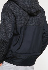 Mörkblå jacka med en kontrasterande, texturerad huva med ett vertikalt grått mönster. Designen inkluderar elastiska ärmslut och fåll.