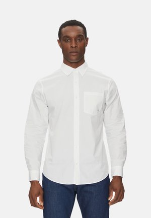 Camisa blanca de manga larga con botones y cuello puntiagudo, que presenta un bolsillo en el lado izquierdo del pecho y una textura suave. Llevada con vaqueros azul oscuro.