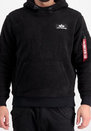 Mężczyzna ubrany w czarną bluzę z polaru z kapturem, z logo Alpha Industries na piersi oraz czerwoną metką "Remove Before Flight" na kieszeni na lewym rękawie.