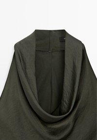 Olivgrön draperad topp i ett mjukt, texturerat tyg. Har en vid halsringning och minimal sömdetaljering som förstärker flödet och elegansen.