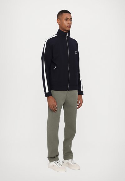 Veste zippée navy avec des rayures blanches sur les côtés, deux poches et un logo, assortie à un pantalon olive et des baskets blanches. Texture lisse.