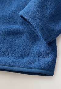 Tessuto in pile blu con una superficie testurizzata. Presenta un orlo cucito e un piccolo dettaglio del logo vicino al polsino in una tonalità più scura.