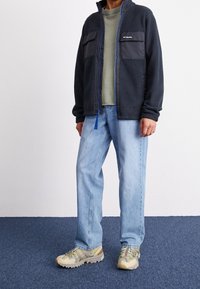 Męska granatowa bluza polarowa z czarnym nylonowym przodem, niebieskim zamkiem błyskawicznym, dwiema kieszeniami na piersi; w komplecie z jasnoniebieskimi dżinsami i beżowo-szarymi butami trekkingowymi.