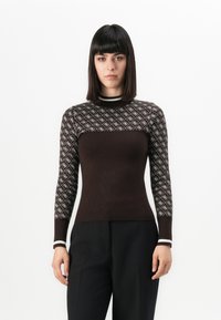 Mujer con cabello oscuro liso, vistiendo un suéter ajustado marrón con mangas estampadas y pantalones negros, de pie contra un fondo blanco.