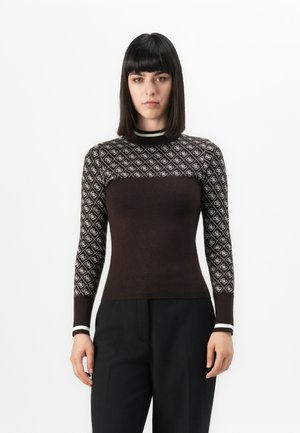 Femme aux cheveux raides et foncés, portant un pull marron ajusté avec des manches à motifs et un pantalon noir, se tenant devant un fond blanc.