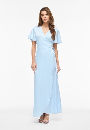 Femeie stă purtând o rochie tip wrap albastru deschis, cu mâneci scurte, ușor evazate și sandale argintii cu vârful deschis, pe un fundal alb simplu.