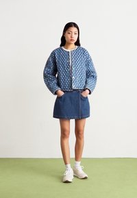 Μπλε τζιν μπουφάν με λουλουδάτο σχέδιο, κοντή σχεδίαση και κουμπί μπροστινό κλείσιμο, συνδυασμένο με τζιν φούστα. Το μοντέλο φοράει λευκά sneakers.