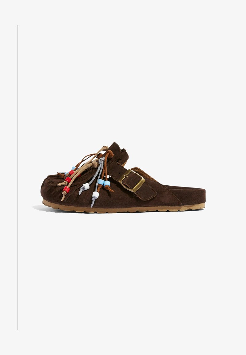 La chaussure slip-on en daim marron présente des détails en franges avec des perles colorées, une semelle plate et une boucle latérale ajustable. Texture lisse sur l'ensemble.