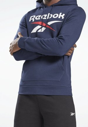 Personne portant un sweat à capuche Reebok bleu marine avec un logo rouge et blanc et un pantalon noir avec un petit logo blanc Reebok, bras croisés sur la poitrine.