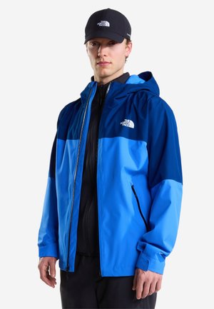 Man draagt een tweekleurige blauwe waterdichte jas met capuchon en een zwarte pet, beide met het The North Face-logo, poseert tegen een effen achtergrond.