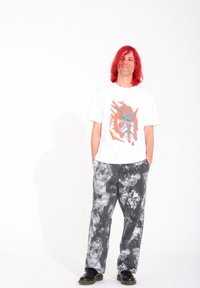Weißes T-Shirt mit einem bunten Grafikdesign, das einen Tiger zeigt, kombiniert mit lockeren, grau gefärbten Hosen im Tie-Dye-Look und schwarzen Schuhen.