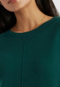 Pull en tricot vert avec un col rond, présentant une texture subtile et une couture centrale sur le devant. Design simple et classique.