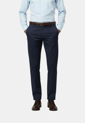 Uomo che indossa pantaloni eleganti blu navy, camicia azzurro chiaro infilata dentro, cintura di cuoio marrone e scarpe eleganti marroni, in piedi con le mani nelle tasche.