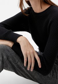 Personne portant un pull noir texturé et un jean gris délavé, assise avec un bras reposant sur un genou plié, sur un fond blanc.