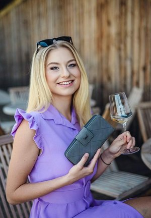 Šedá prošívaná peněženka s klopou a logem, držena v jedné ruce; druhá ruka drží sklenici bílého vína. Pozadí má dřevěné dekorace.