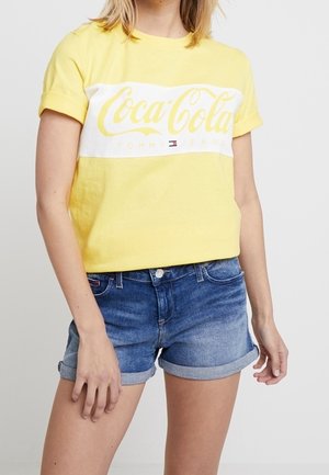 Yellow cotton t-shirt with a white horizontal band displaying the Coca-Cola logo. Paired with rolled-up denim shorts.