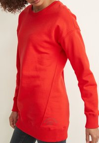 Femme portant un sweat-shirt à manches longues rouge vif avec des poignets et un ourlet côtelés, associé à un jean foncé, devant un fond uni.