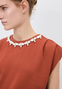 Top v rezavé barvě s kulatým výstřihem ozdobeným bílým perlovým náhrdelníkem. Hladká látka s jemnou texturou a krátkými rukávy.