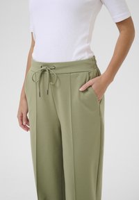 Pantalons en tissu doux de couleur vert olive avec taille à cordon, poches latérales et coupe décontractée. Présente des coutures verticales pour un détail supplémentaire.